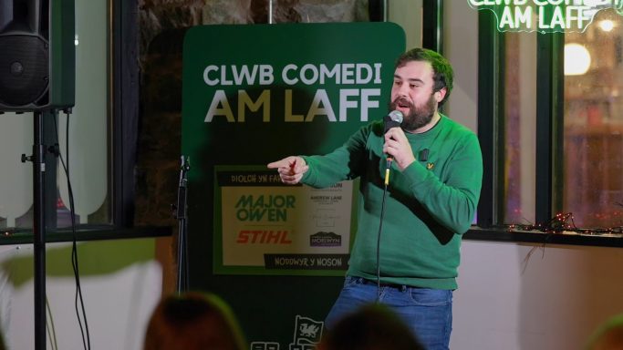 A man in a green sweater performs comedy on stage, pointing to the audience.