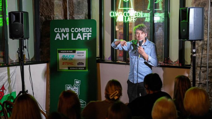Stand-up comedian performing on stage with an audience seated in the foreground.